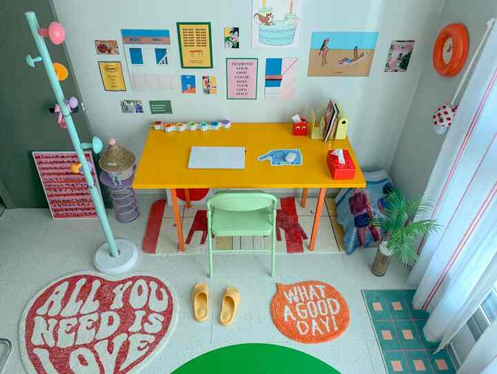 黄色いデスクとライトグリーンの椅子が中心にある、カラフルなポスターの壁と複数のラグが置かれた明るく活気のあるワークスペース