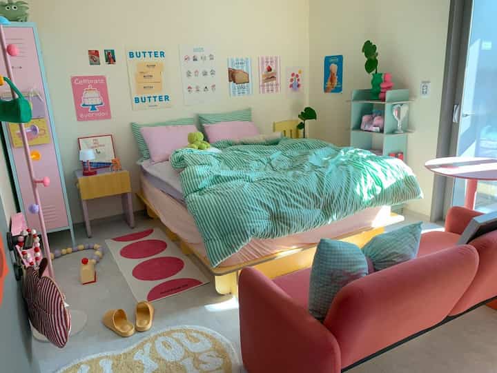 明るいナチュラルトーンにピンクとグリーンが調和した二人暮らし向けの寝室、ベッドとソファ、壁のポスターが配置された雰囲気のあるインテリア