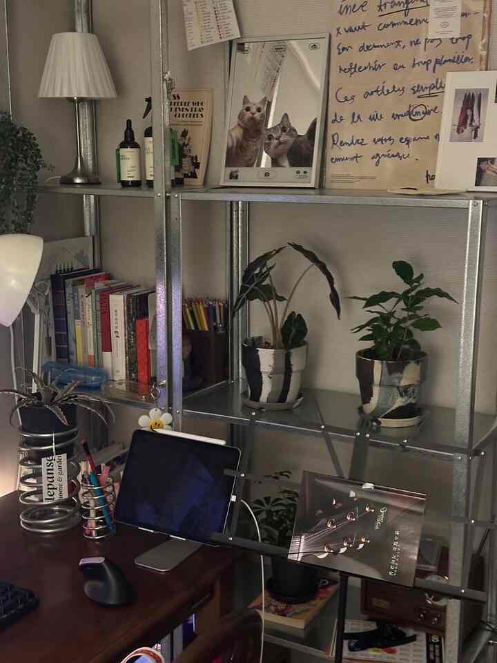 温かみのある木製デスクと金属製の棚が配置されたホームオフィス空間で、植物や本、タブレットスタンドと照明がある居心地の良い作業環境