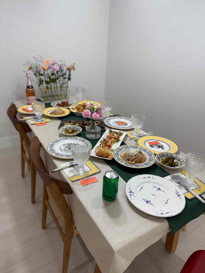 ベージュのテーブルクロスとグリーンのテーブルランナーが配置されたキッチンダイニング、ヴィンテージの食器、花瓶、多彩な料理が飾られたホームパーティーのインテリア