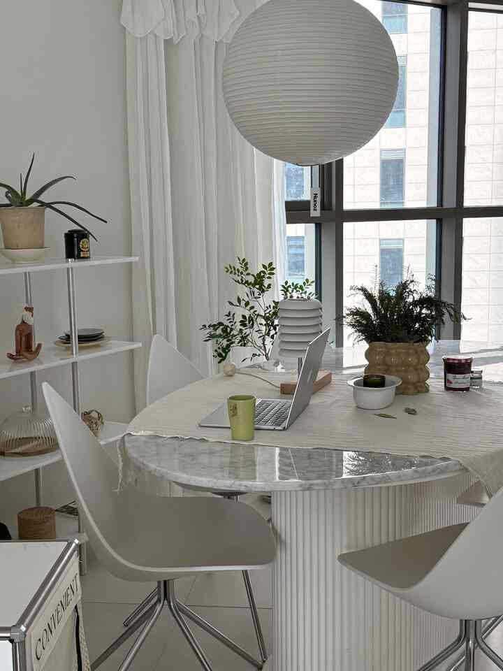 明るいホワイトトーンのキッチンダイニング空間、丸いダイニングテーブルとテーブルクロス、白いダイニングチェア、ペンダントライト、植物が配置されたナチュラルモダンスタイルのインテリア