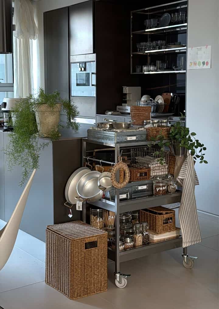 ホワイトとグレーのトーンで構成されたキッチン、中央に金属製のキッチンワゴンとラタンバスケット、緑の植物が配置された印象的なインテリア