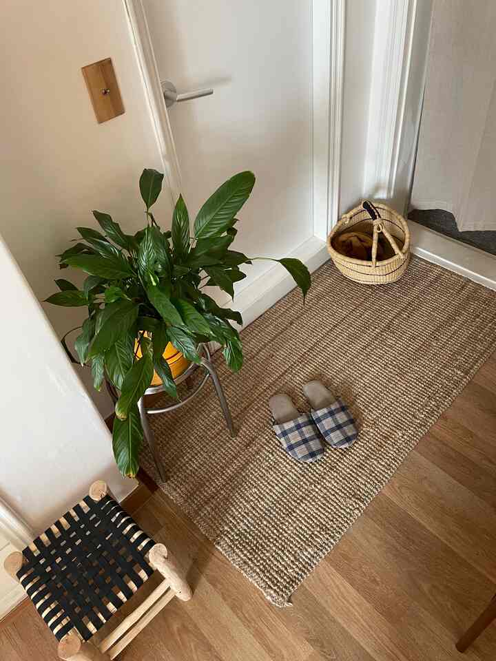 ナチュラルな色調の狭い玄関、黄麻のラグと木製スツール、緑の植物、チェック柄のスリッパ、バスケットが配置された温かみのあるインテリア
