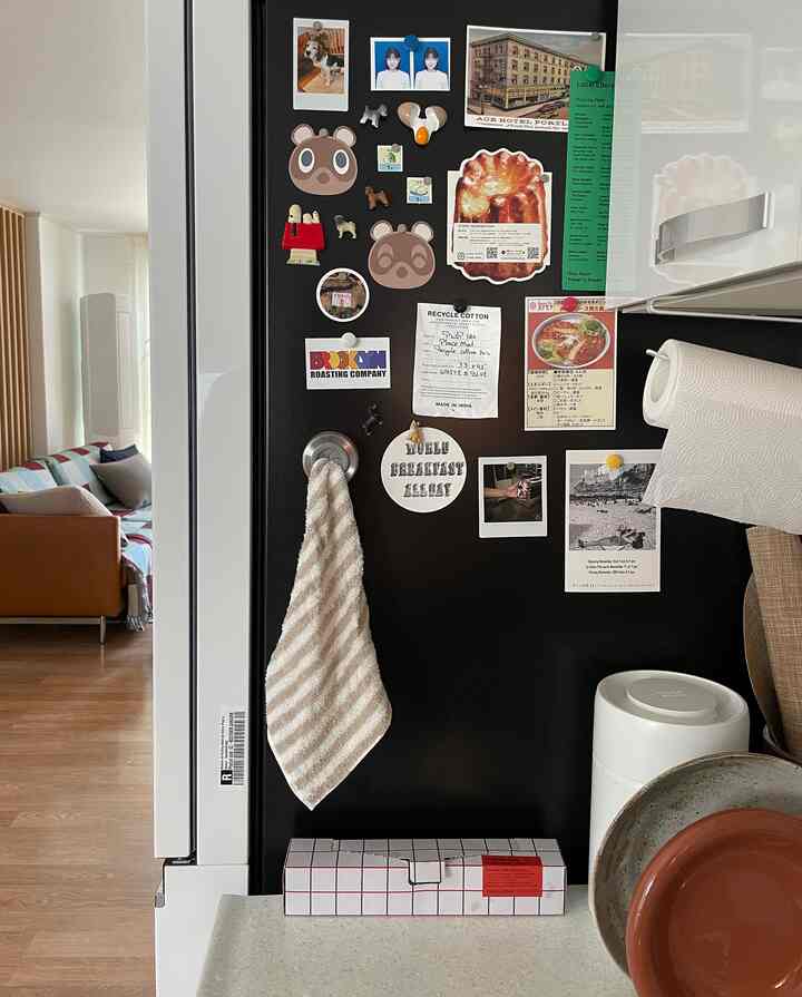 黒の冷蔵庫側面に様々な飾りが配置され、左側にブラウンのソファが見える暖かいリビング空間