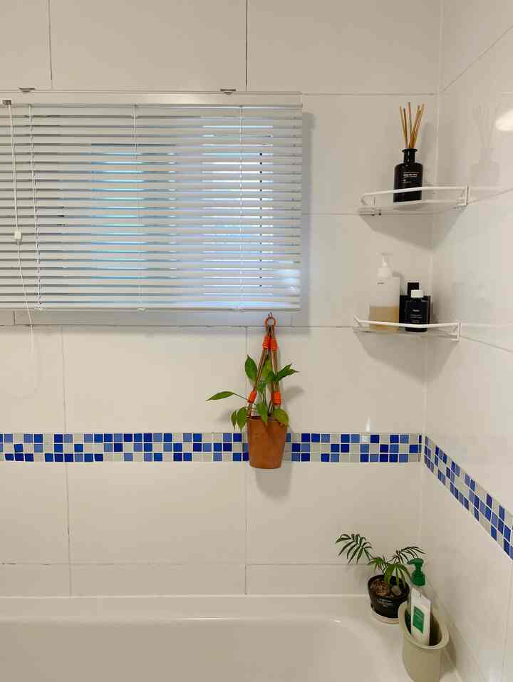白いタイルと青のモザイクが調和するバスルーム、白いブラインドとコーナー棚が清潔感のあるインテリアを演出