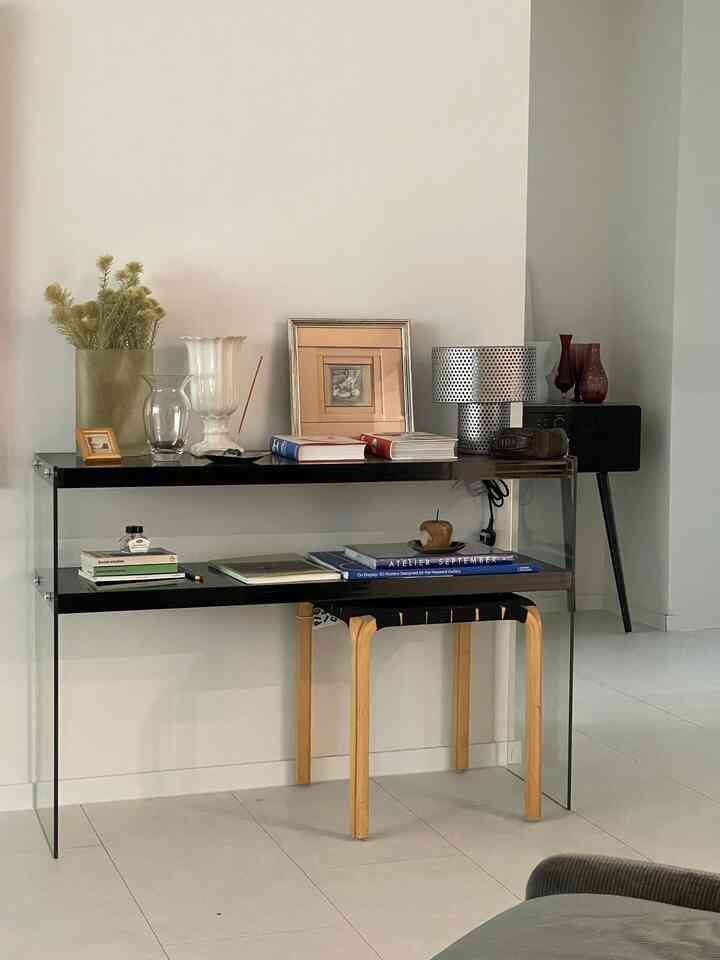 明るいホワイトトーンの壁とタイル床に、透明なガラス製の本棚が設置され、本や額縁、ランプが配置されたミニマルな廊下の空間