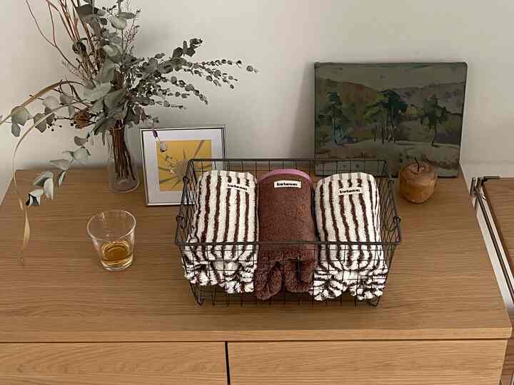 ナチュラルウッド調の寝室チェストの上にドライフラワーの花瓶、折りたたまれたタオル、額縁とオブジェが配置された雰囲気のあるインテリア