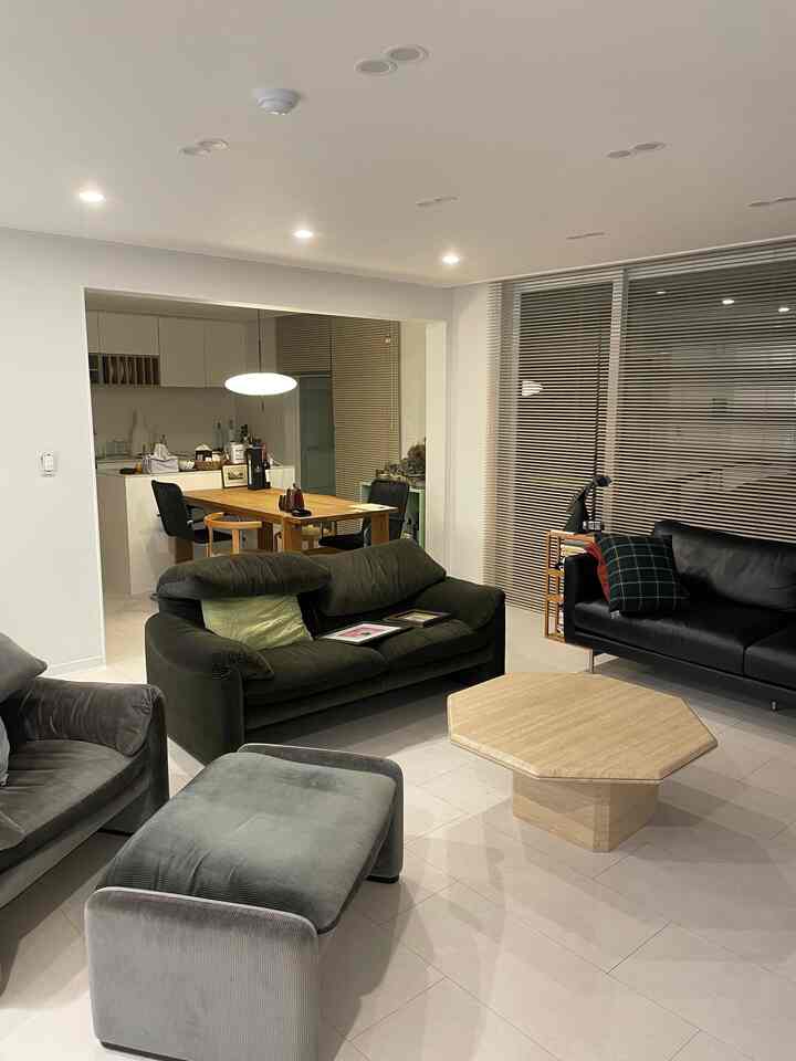 ナチュラルカラーのモダンなリビング、2つのソファと六角形のコーヒーテーブル、背景に木製ダイニングテーブルとブラインドが配された雰囲気のあるインテリア