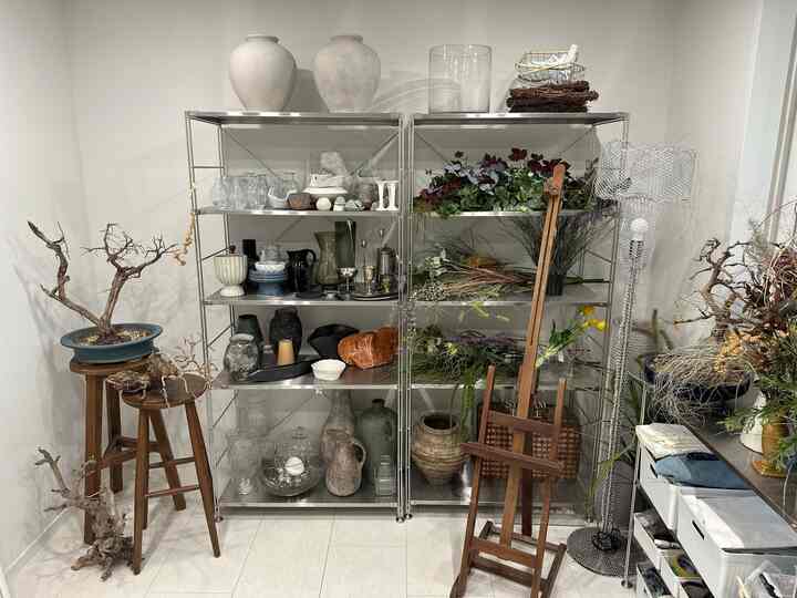 白い壁と床に茶色の木製スツール、メタル棚、多様な植物とオブジェが配された自然な雰囲気の収納空間