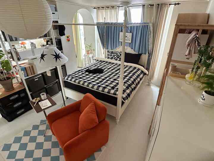 落ち着いた木目調とブルーの遮光カーテンが調和したワンルームの寝室、オレンジのアームチェアとチェック柄の寝具がアクセントの雰囲気あるインテリア