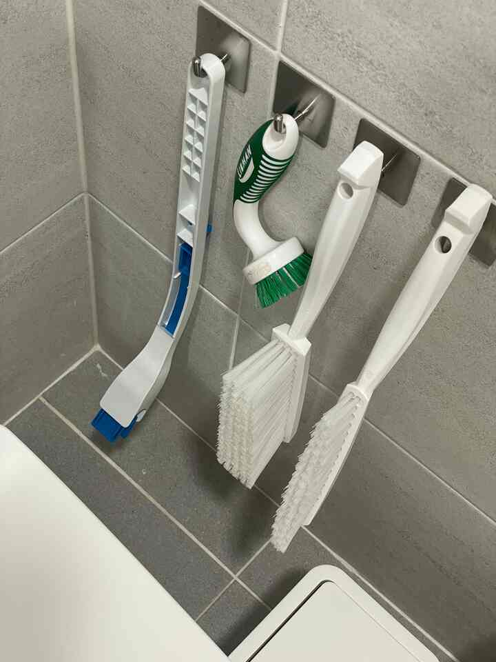 グレーのタイル壁にステンレスフックがあり、浴室の掃除用ブラシと道具が掛けられたミニマルな浴室の一角