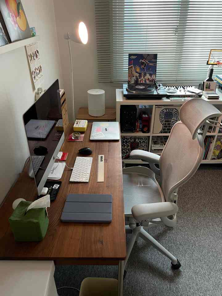 ナチュラルな木目調のデスクとグレーのエアロンチェア、ブラインド、書棚が配置されたモダンなホームオフィスのインテリア