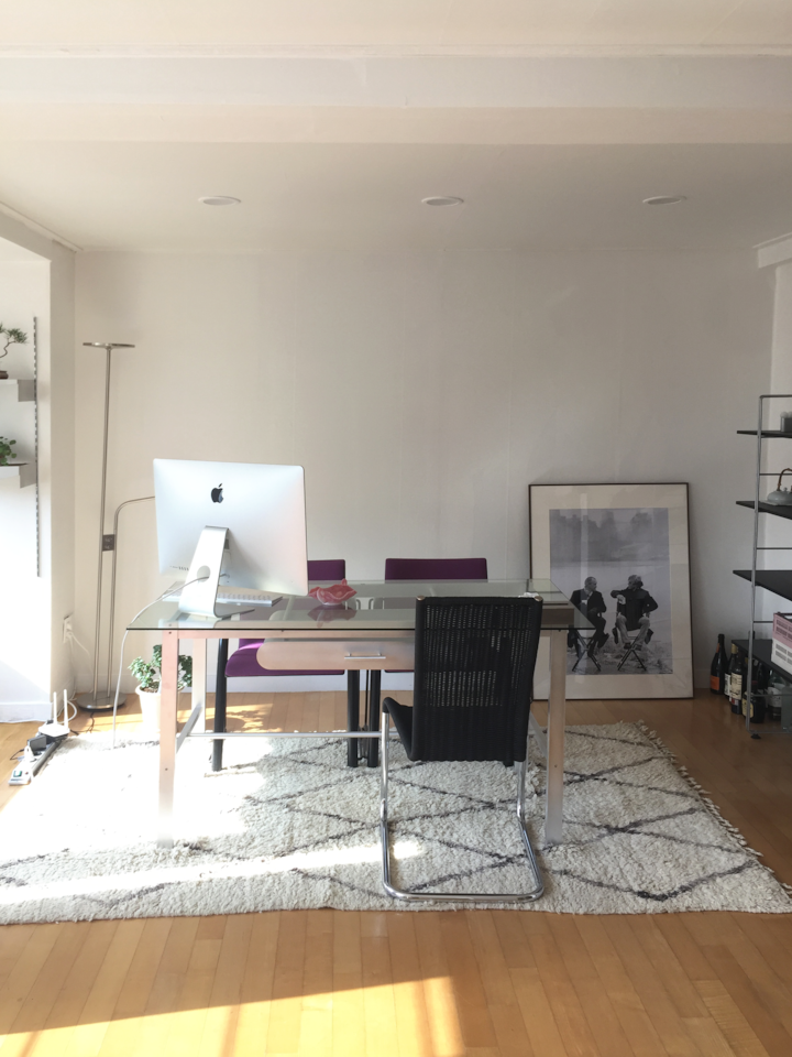 白とシルバーの色調のリビングルームのホームオフィス、透明なガラステーブルと黒のオフィスチェア、クリーム色のラグが配置された雰囲気のあるインテリア