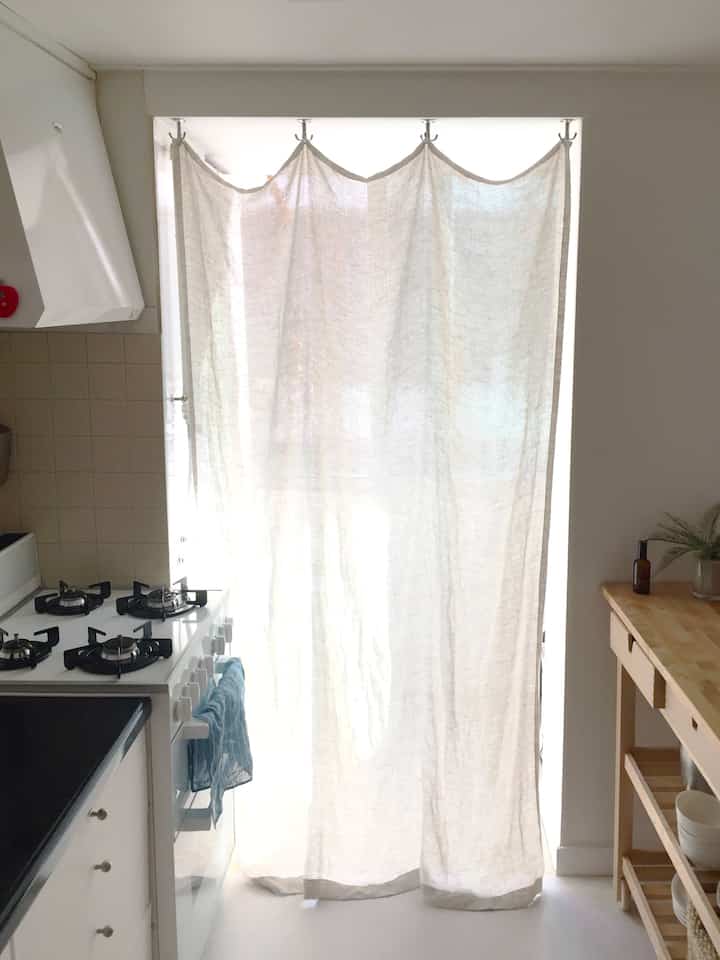 明るいホワイトとウッドトーンが調和したキッチン、リネンカーテンが収納スペースとして使われるナチュラルな雰囲気のインテリア