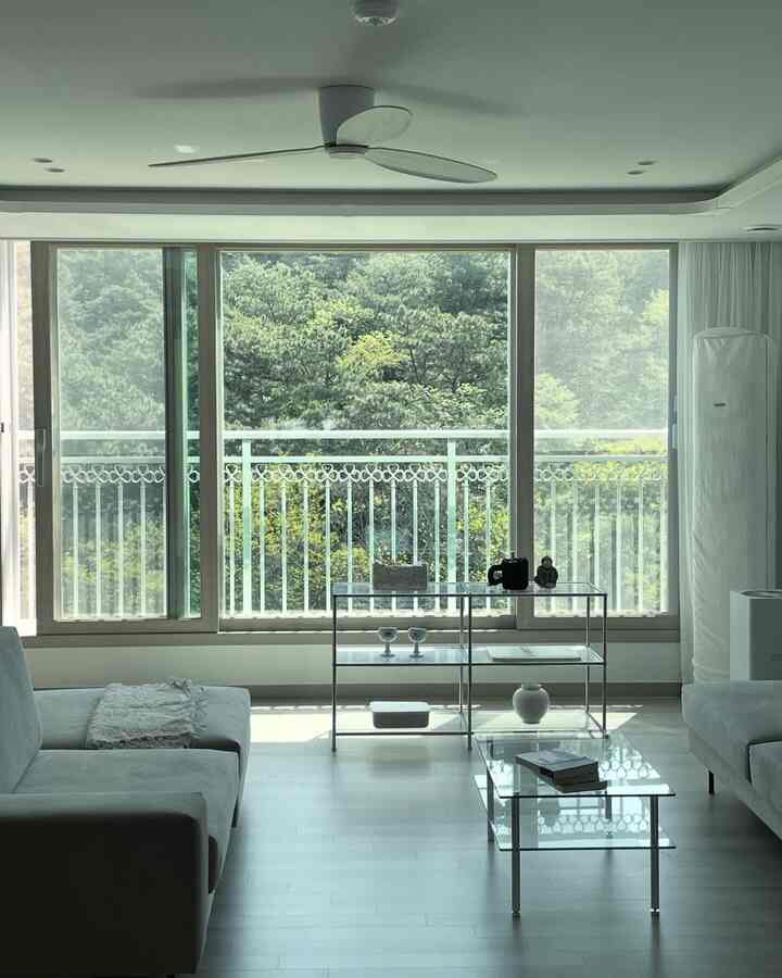 白とグレーのトーンのモダンでシンプルなリビング、大きな窓とカーテン、ガラステーブルが配置された明るく開放的なインテリア