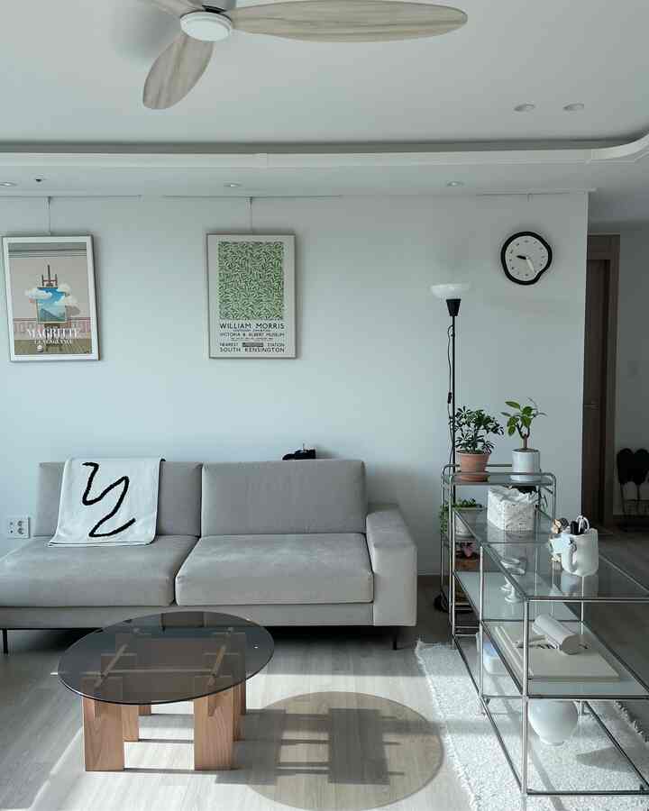 白とグレーの色調のリビング、グレーのソファと丸いガラステーブル、植物とポスターが配置された雰囲気のある空間