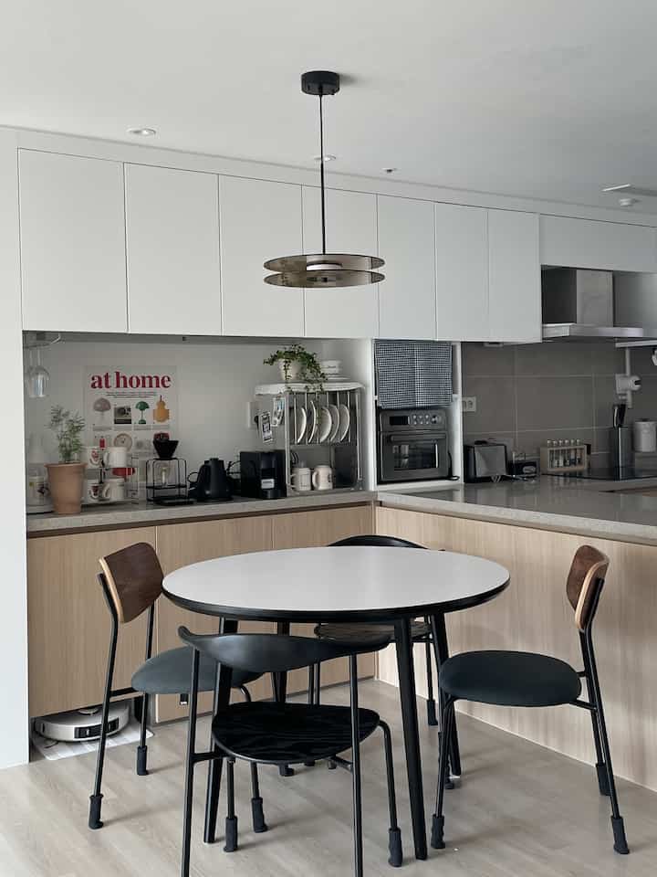 ホワイトの上部キャビネットとナチュラルな木目調の下部収納が調和したキッチン、中央に黒脚の丸いダイニングテーブルが配置された清潔感のある空間