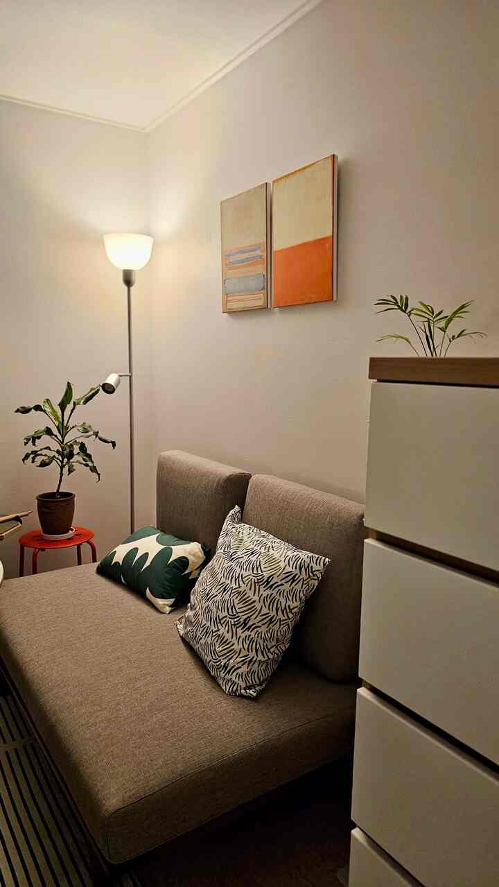 ナチュラルなベージュ調のワンルームリビング、ベージュのソファとクッション、フロアランプ、植物、壁のアートが配置された落ち着いた空間