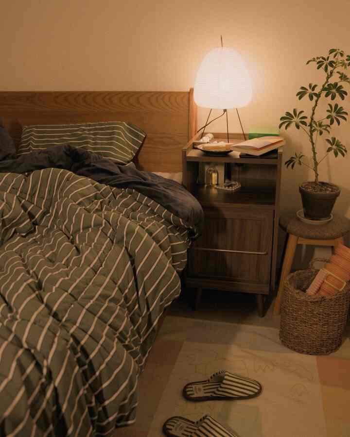 木目調とグリーンのストライプ寝具がある暖かみのある寝室、ナイトテーブルのテーブルランプと植物が配置された居心地の良い空間