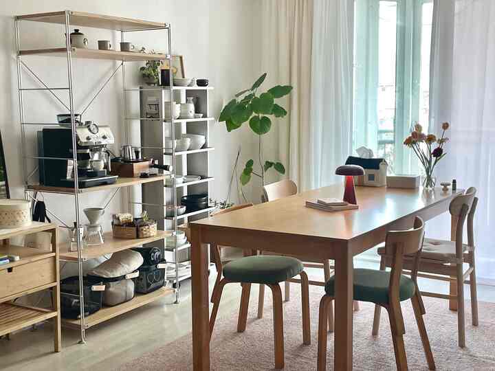 明るくナチュラルな色調のダイニングルーム、木製のテーブルと椅子、コーヒーマシンと食器棚、窓際の観葉植物が配置された雰囲気の良い空間