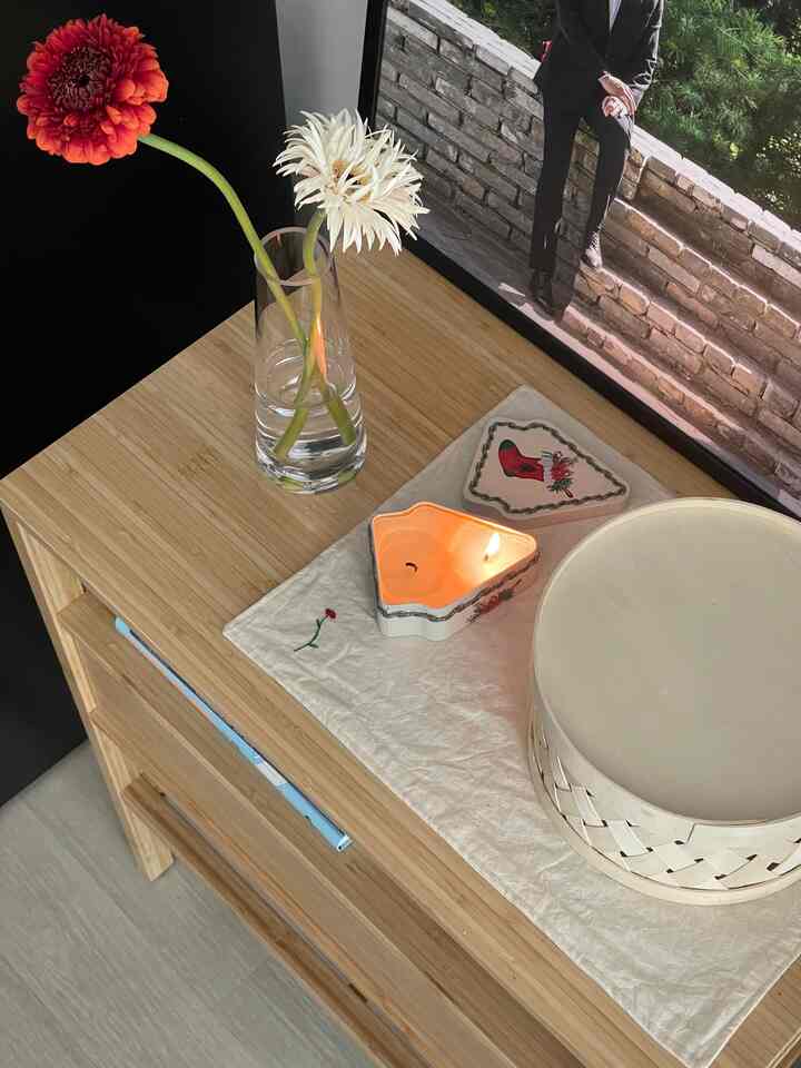 ナチュラルカラーの寝室用ナイトテーブル、花の入った花瓶と灯されたムードキャンドル、写真額が置かれた雰囲気のあるインテリア