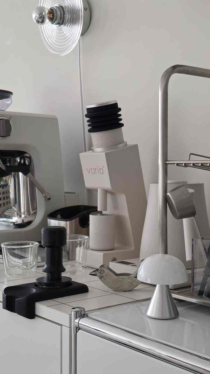 ホワイトのミニマルなキッチン、エスプレッソマシンとグラインダー、テーブルランプが配置された雰囲気のあるおうちカフェ空間