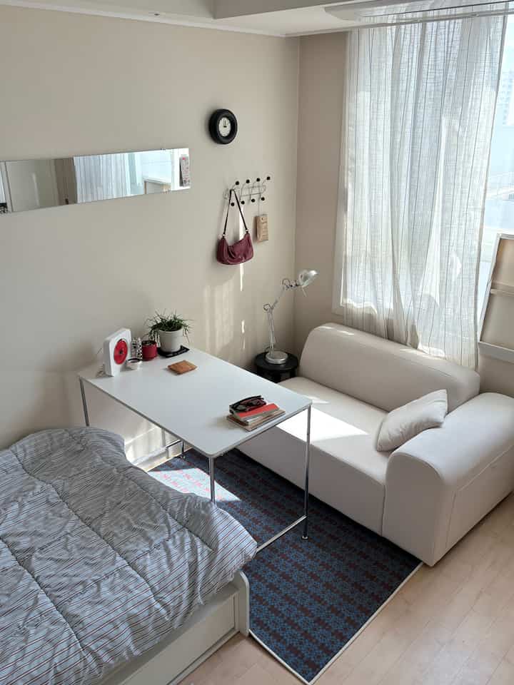 ナチュラルカラーの小さな寝室、窓辺に白いソファ、デスク、ベッドが配置された落ち着いた空間