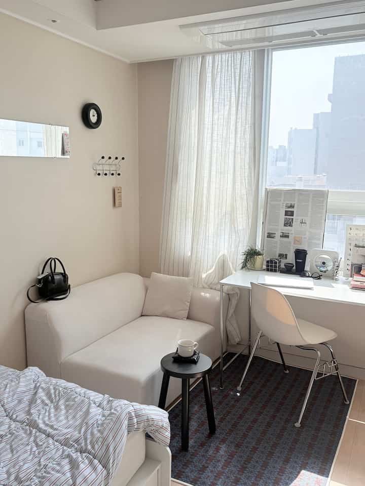 白とダークブラウンの色調のワンルームリビング、ソファとデスクが配置されたシンプルで明るい雰囲気のインテリア