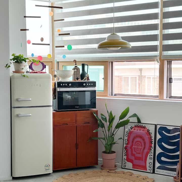 ナチュラルな木目調とクリーム色の冷蔵庫が並ぶコンパクトなキッチンスペース、明るい大きな窓と植物が温かみのある雰囲気を演出