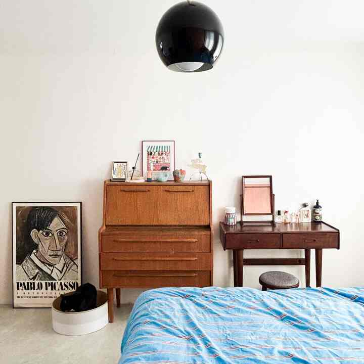 白い壁と床の寝室、木製家具、黒色のペンダントライト、ピカソの絵画、青いベッドカバーのベッドが配置された印象的な空間