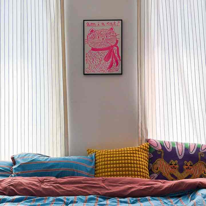 白い壁とカーテンのあるアットホームな寝室、中央に猫のイラストが飾られ、多彩なクッションと青のストライプベッドリネンが配置された温かみのあるインテリア