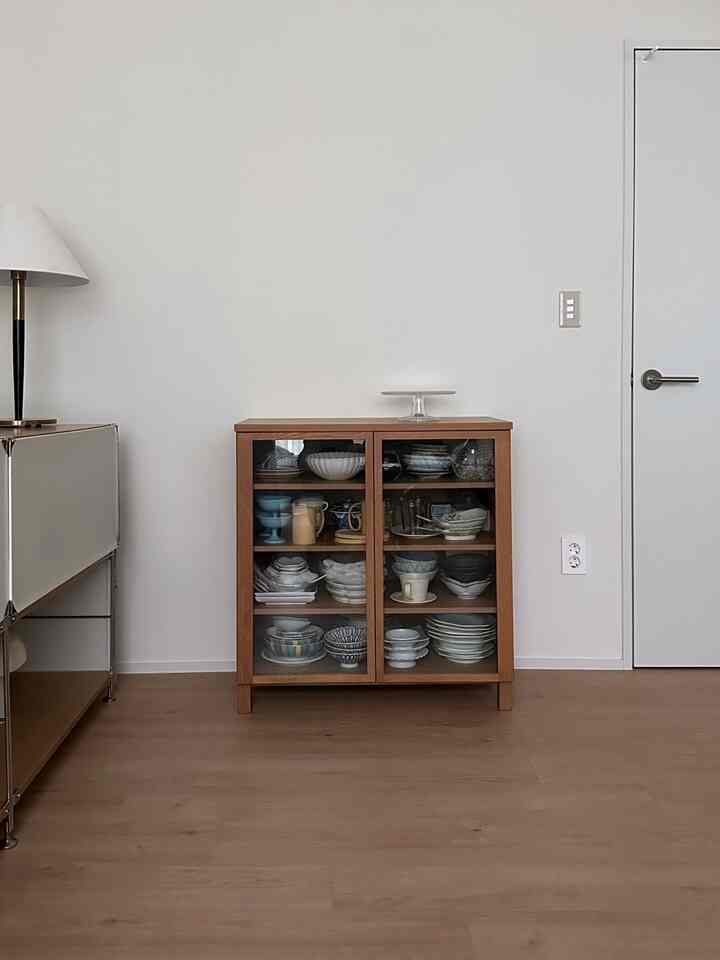 白い壁とウッドトーンの床が調和する空間の中央に木製ガラス扉のキャビネットが配置されたすっきりとした収納インテリア