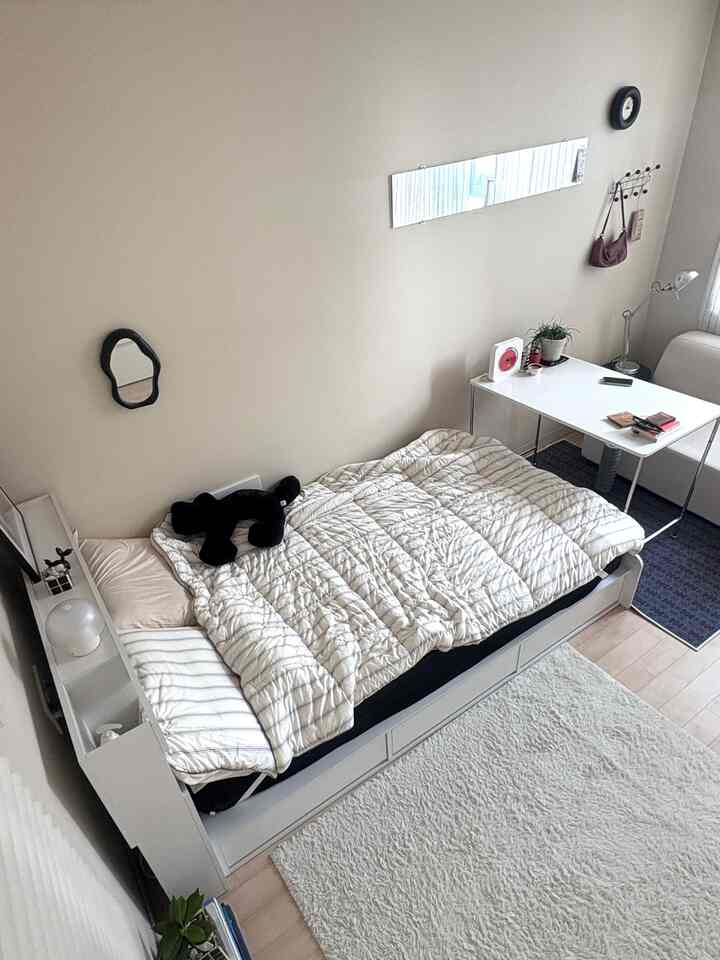 白とナチュラルカラーのワンルーム寝室、ベッドとデスクが配置された落ち着いた清潔感のある空間