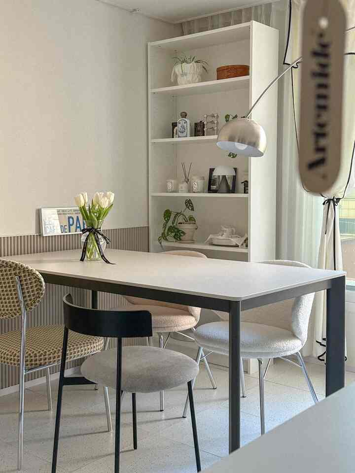 白色のトーンで統一されたリビング、中央に大きなダイニングテーブルと様々なデザインの椅子が配置され、右側に本棚とシルバーのフロアランプがあるモダンなインテリア