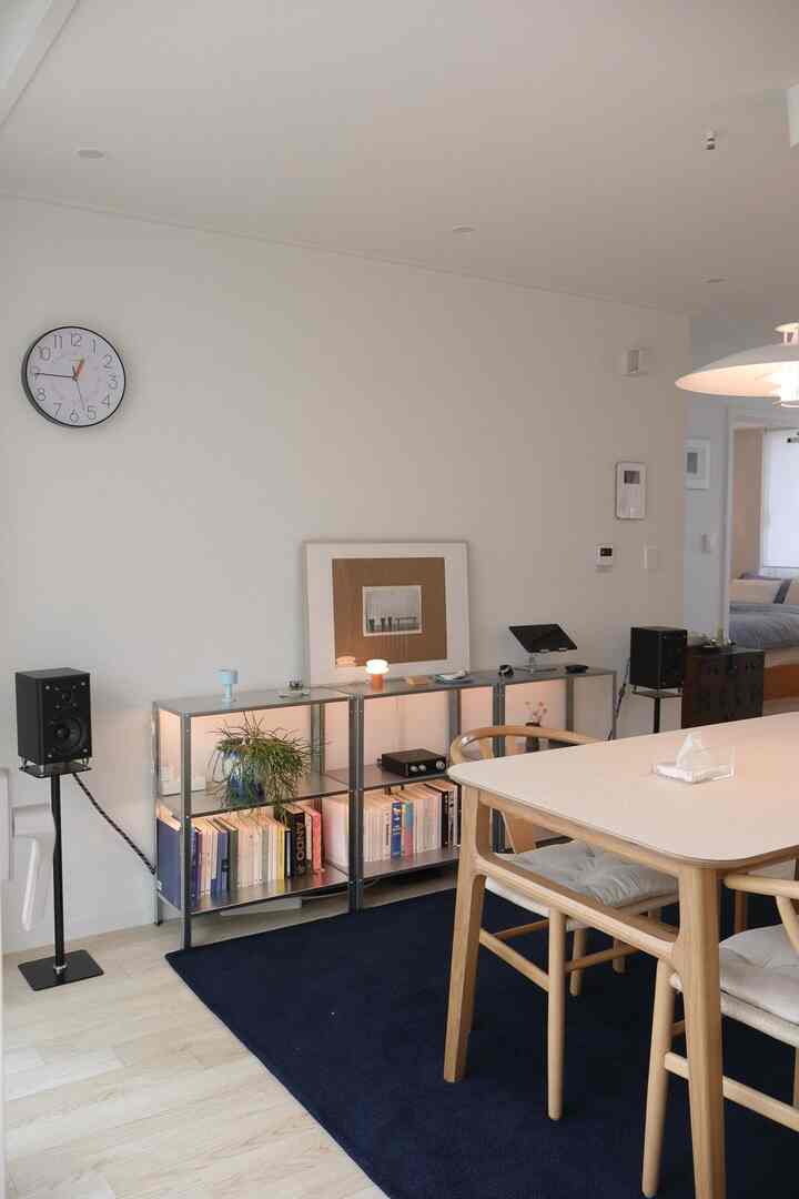 白い壁と木目調のダイニングチェアが配置されたリビング、ネイビーのラグとモダンな棚に本とスピーカーが並ぶ清潔感のある読書と音楽の空間