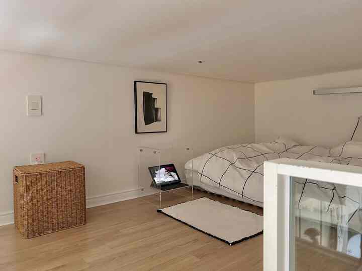 ナチュラルな木目調の床と白い壁のワンルーム寝室、透明なアクリルのナイトテーブルとシンプルでモダンな幾何学模様の寝具が配置された空間