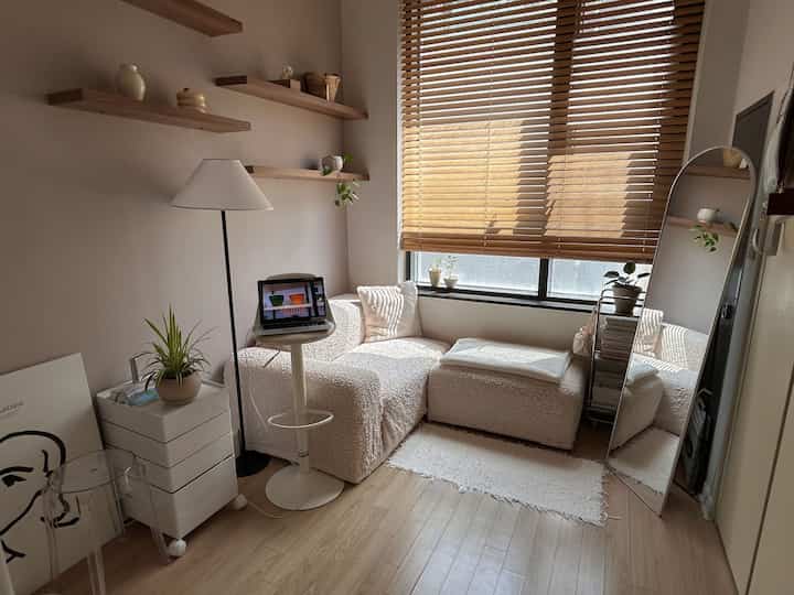 ナチュラルなトーンの狭いリビング、シンプルな白いソファと木製ブラインド、全身鏡とランプが配置された落ち着いた空間