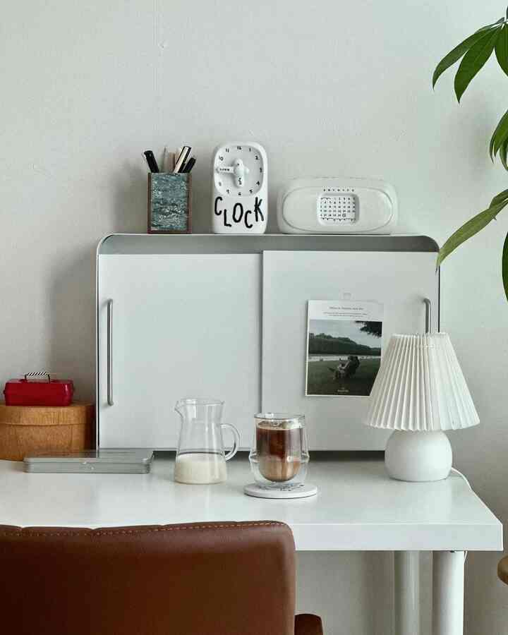 明るいホワイトトーンの小さな部屋のデスクスペース、白いスリム収納キャビネットと整ったホームカフェの小物が配置されたシンプルなインテリア