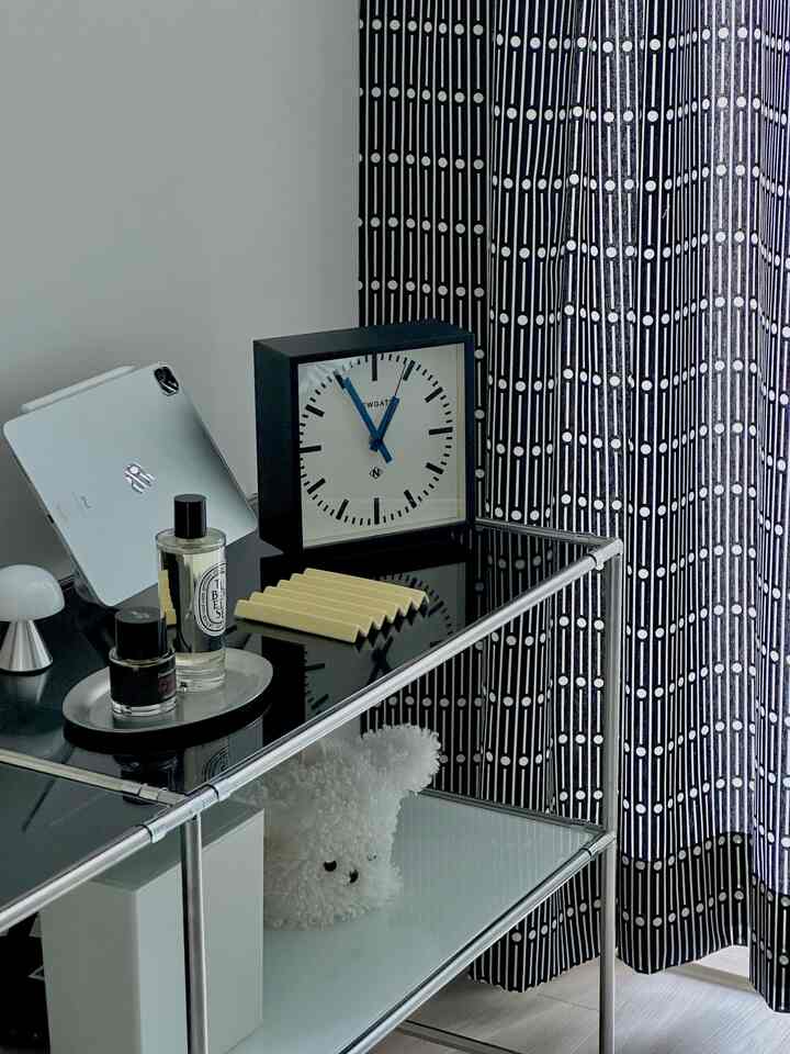 モダンで清潔感のある小さな部屋、金属製の収納棚に置かれた時計と香水、黒白パターンのカーテンのそばの空間