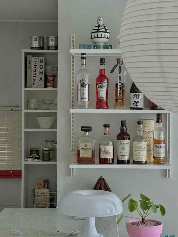 白を基調としたキッチンの壁に設置された棚に様々なウイスキーの瓶とオブジェが置かれた、清潔で実用的なホームバー空間
