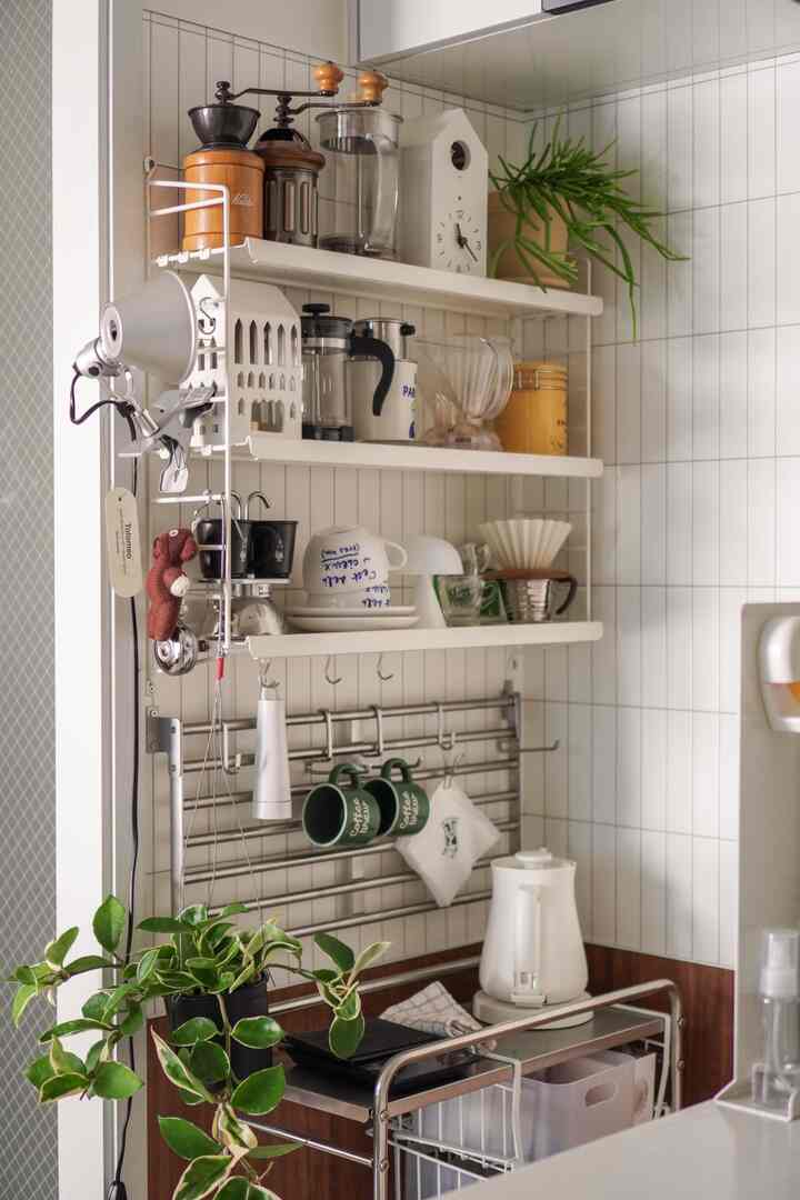 白とステンレスの調和が取れたキッチン空間、壁の棚や電気ケトルなどホームカフェ要素が配置された清潔感のあるインテリア