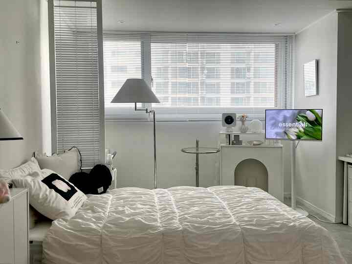 ホワイトトーンの寝室、清潔な寝具とモダンなフロアランプ、デスクとモニターが配置されたシンプルな空間