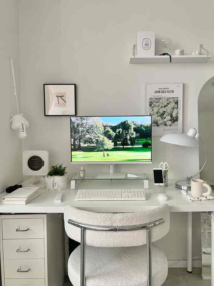 明るいホワイトトーンの寝室に配置されたホームオフィス、中央に白いデスクとモニタースタンド、シンプルモダンな家具が配される印象的な空間