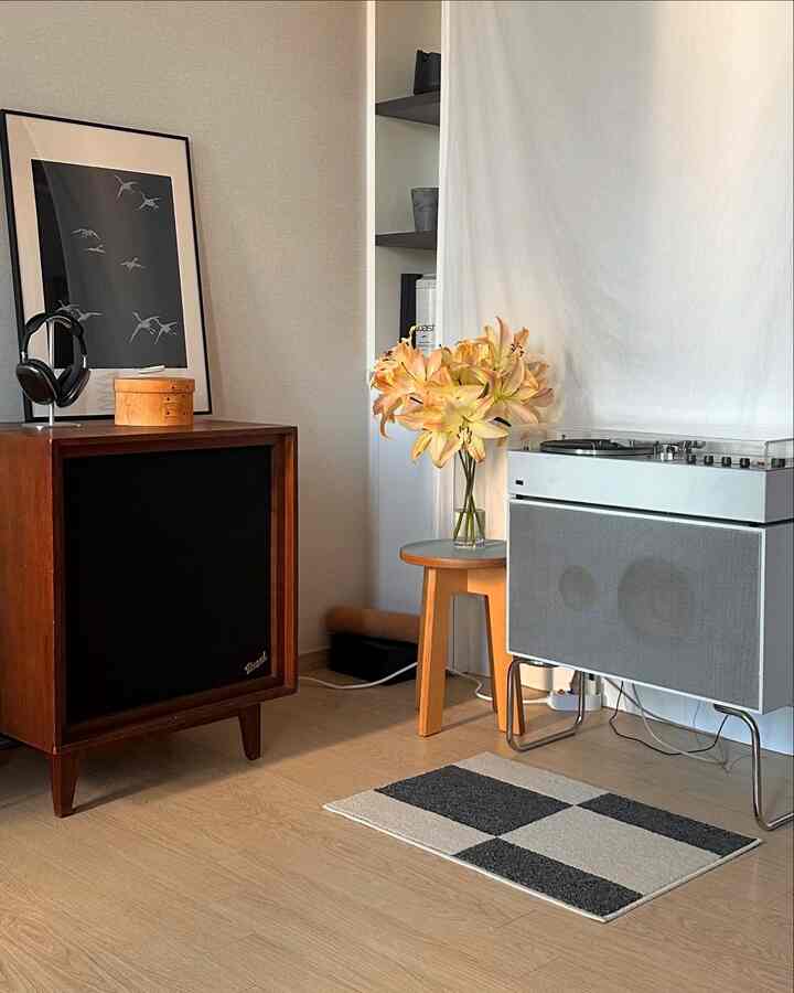 ナチュラルなブラウン調のリビング空間、ブラウンのスピーカーとターンテーブル、木製スツールの上に花瓶と黄色い花が配置された雰囲気のあるヴィンテージインテリア