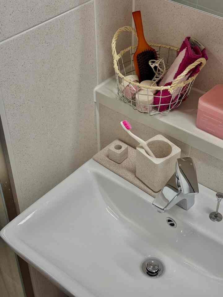 ベージュのタイル壁と白い洗面台が配置された清潔感のあるバスルーム、ピンクのタオルと歯ブラシがある実用的な収納スペース