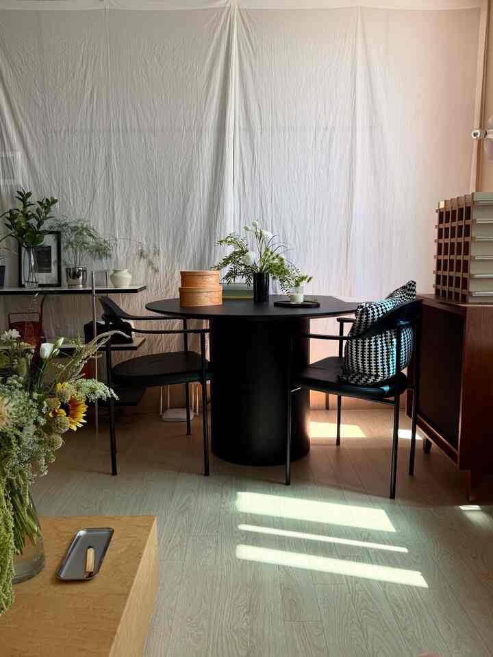 ナチュラルな木目調の床とベージュのカーテンを背景に、中央に黒の丸テーブルと椅子が配置された落ち着いたダイニングルーム
