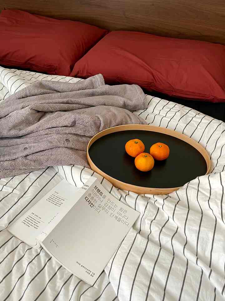 ナチュラルカラーの寝室、ストライプの掛け布団カバーと赤い枕、トレイにみかんが置かれた落ち着いた読書空間