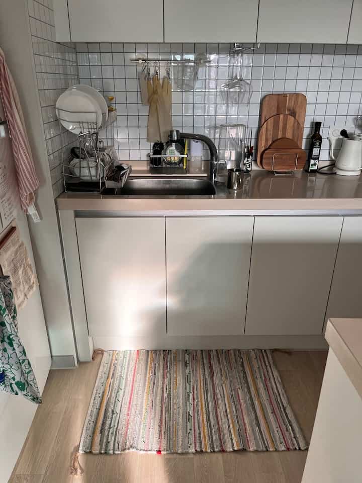 ホワイトとナチュラルカラーのキッチンスペース、シンク周りの調理器具と整然としたラグが配置された実用的なインテリア