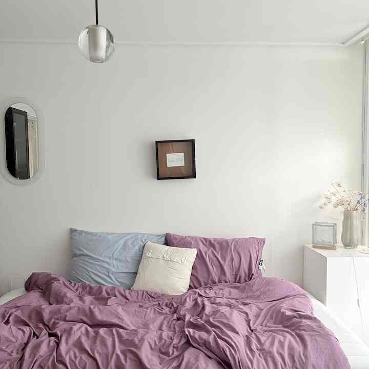 白い壁と自然光が差し込む、ラベンダーカラーの寝具が置かれたシンプルな寝室