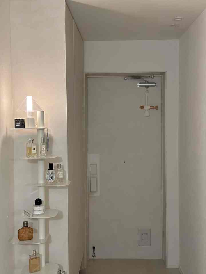 明るいホワイトトーンの玄関スペース、扉の横に白い棚があり香水と照明が飾られた印象的な空間